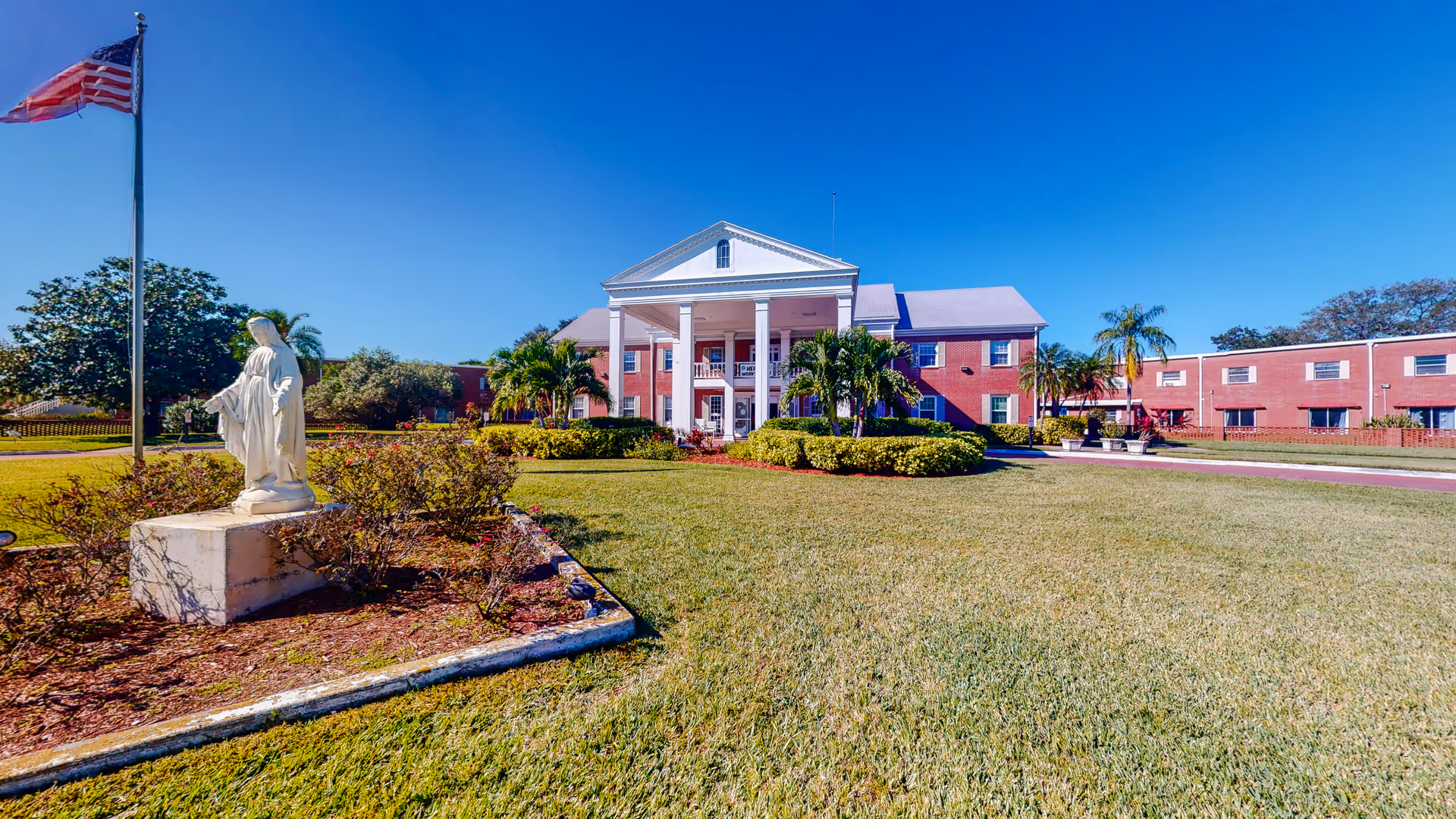 Front lawn of a brick senior living facility with statue, flagpole, and palm trees.