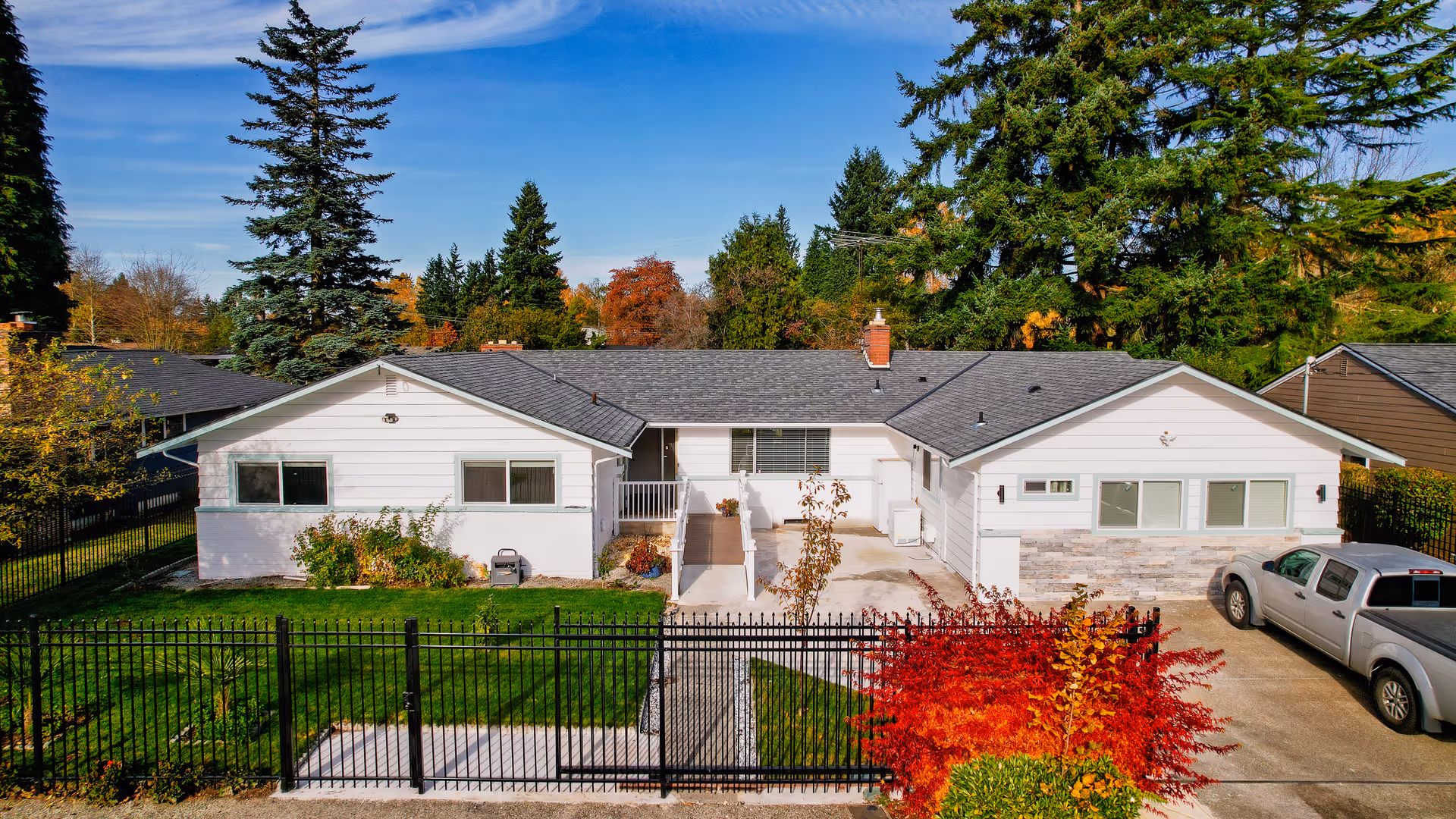 Single-story white senior living facility with a black fence, driveway, and autumn trees.