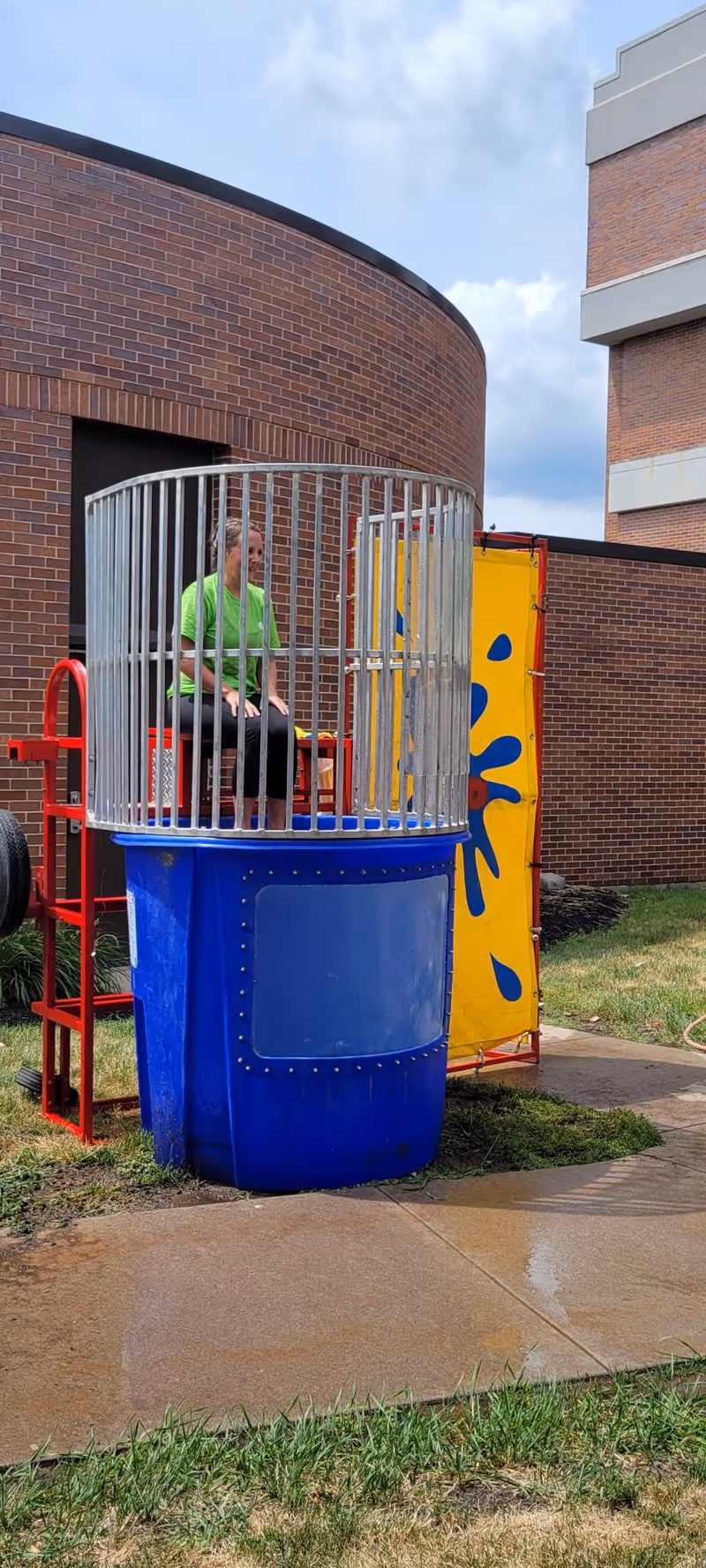 A person sits on the seat of a dunk tank set up outdoors in front of a brick building.