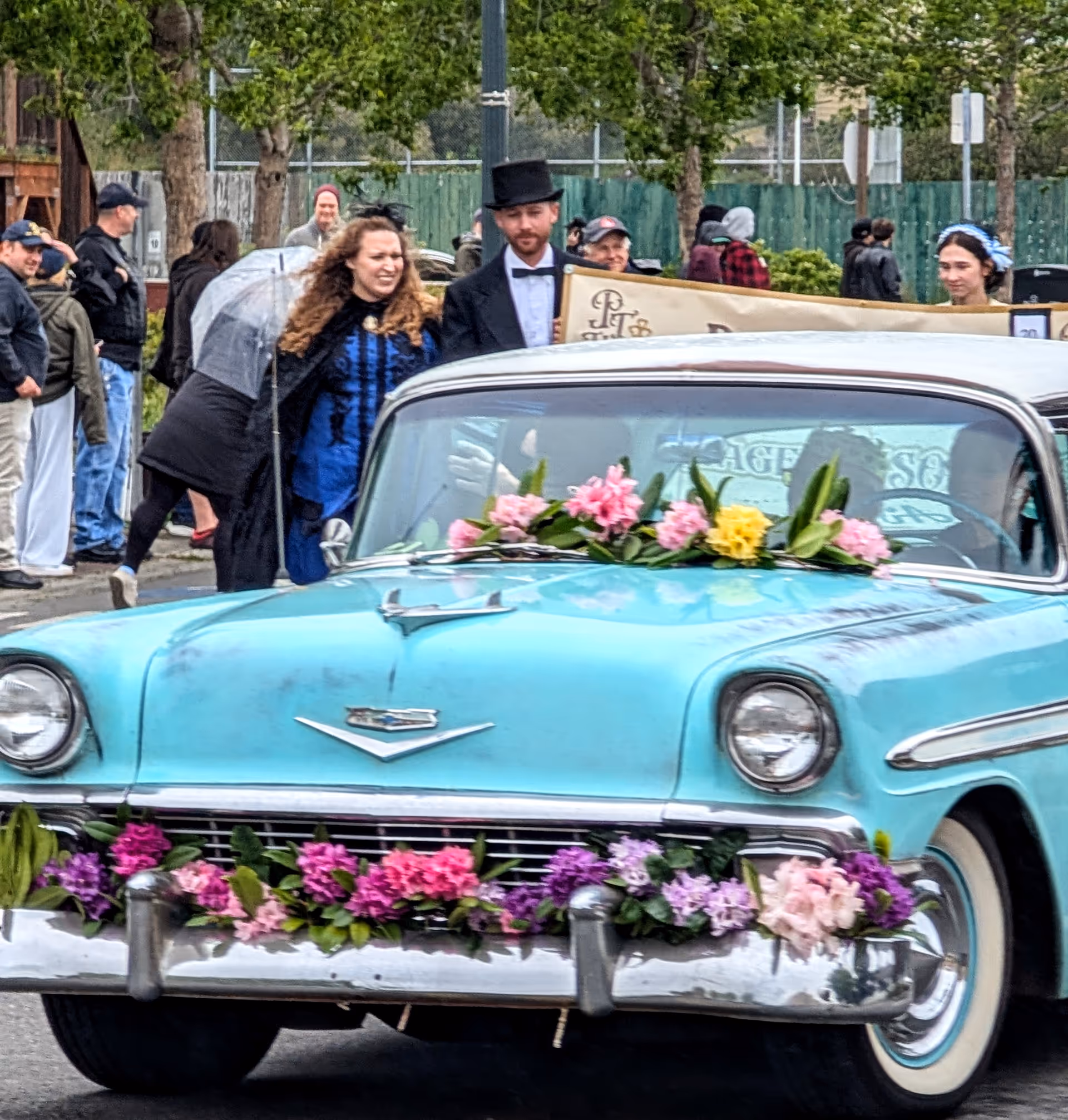 A vintage turquoise car decorated with flowers on a street, with people in background.
