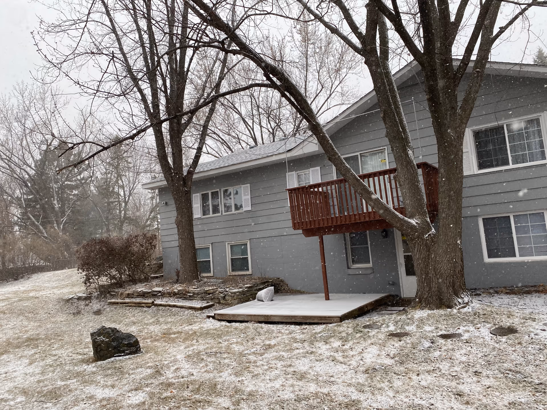 Two-story gray house with a small red balcony, large trees, and a light dusting of snow on the yard.