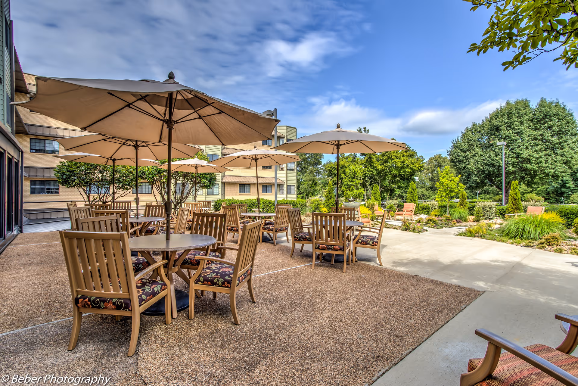Outdoor patio with wooden tables, cushioned chairs, and large umbrellas in a landscaped courtyard.