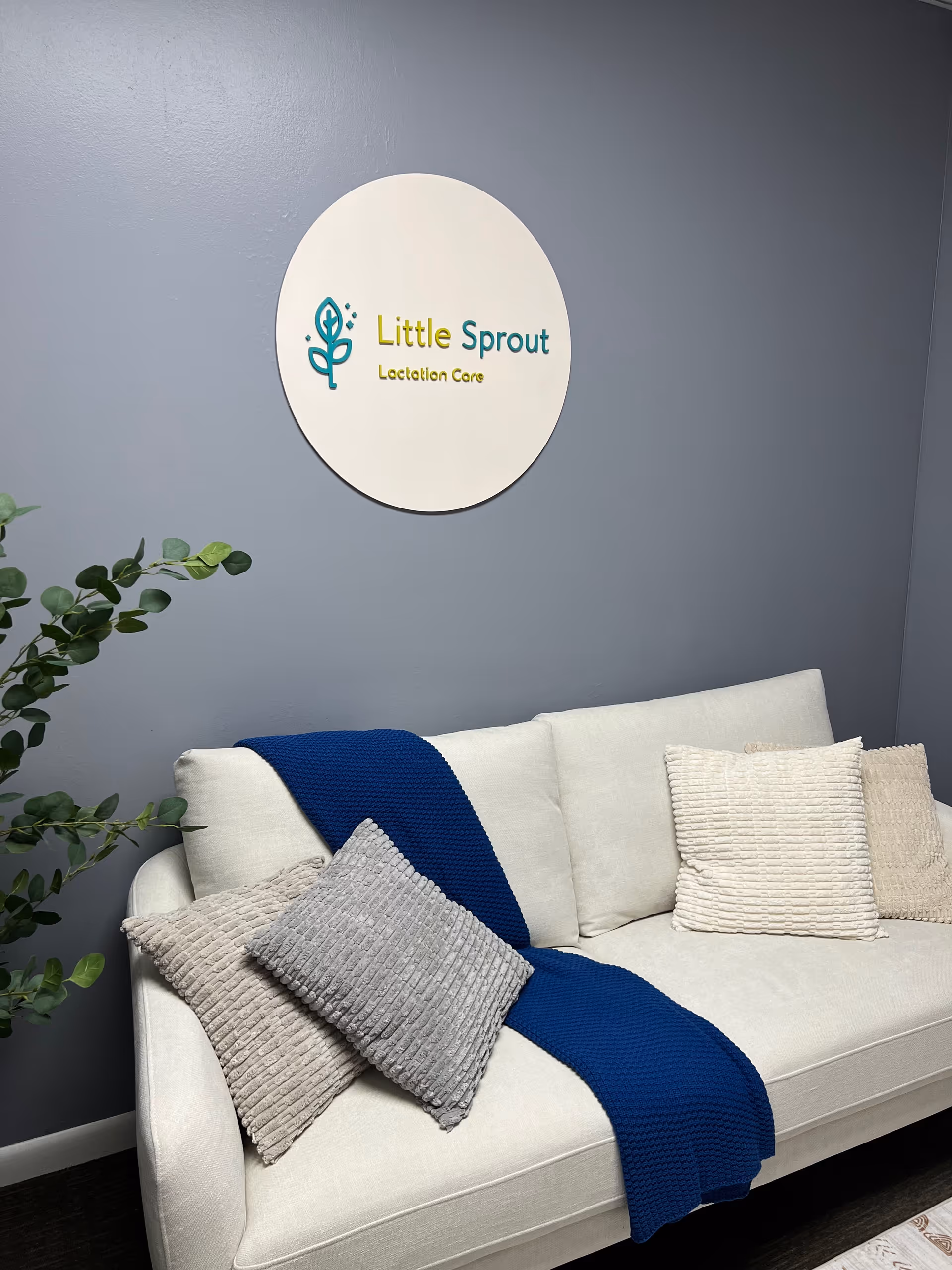 Lounge area featuring circular Little Sprout sign, blue-gray wall, and white sofa with blue throw.