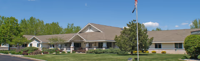 One-story senior living building with a landscaped lawn, flagpole, and clear blue sky.