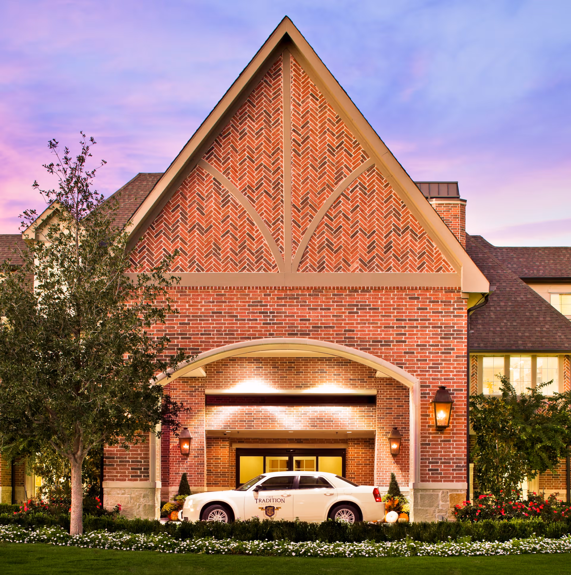 Front exterior view of a senior living facility named The Tradition–Prestonwood during sunset, featuring a large brick building with a steep gabled roof, well-maintained landscaping with trees and flowers, and a white car parked under the entrance canopy.