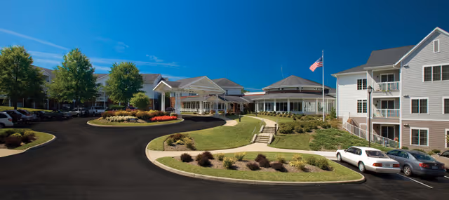 Wide exterior view of Brandon Oaks Life Plan Community featuring a large driveway with parked cars, landscaped greenery, and multiple connected buildings under a clear blue sky with an American flag on a flagpole.