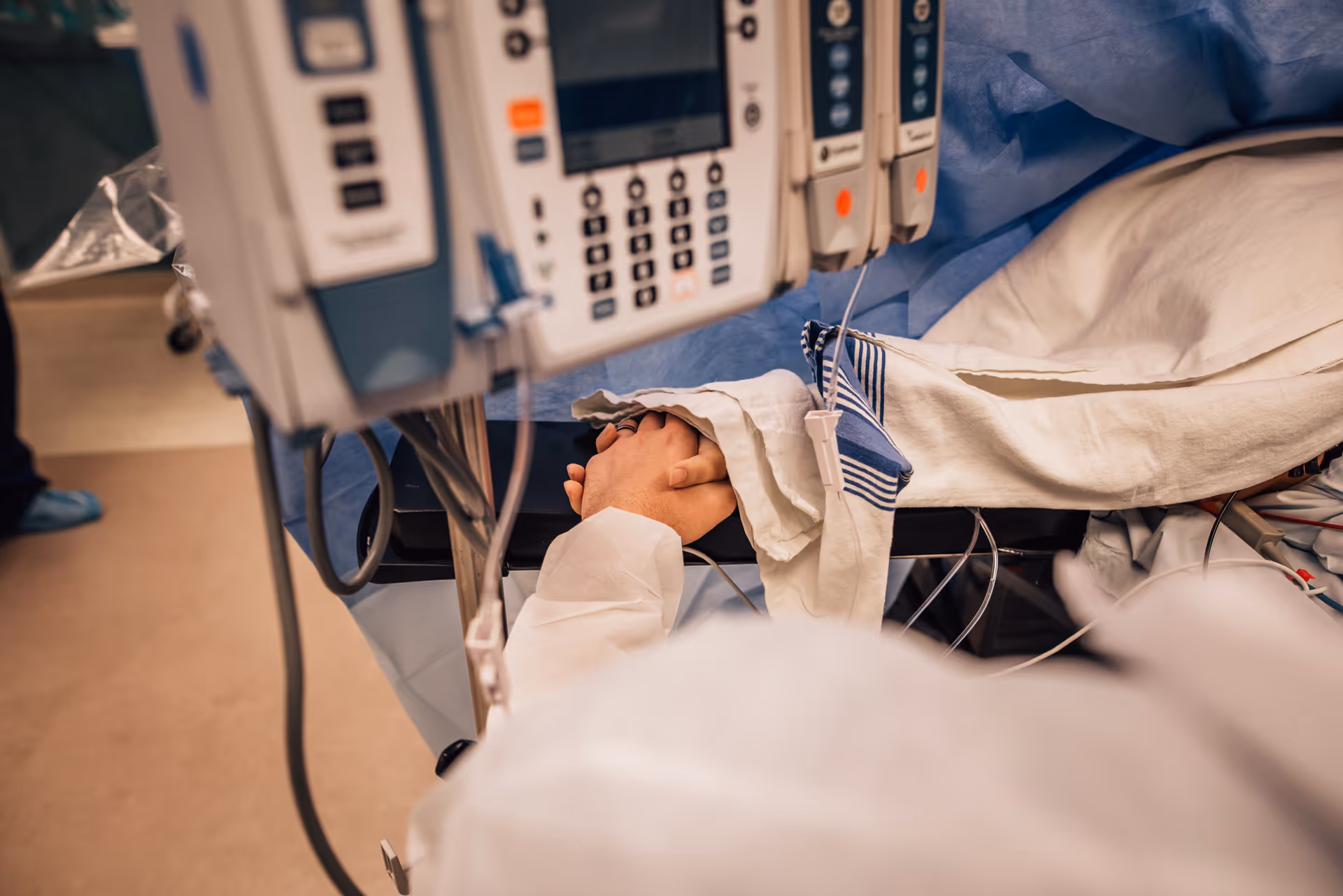 Medical bed with IV lines and monitors, hands clasped beneath sheets in hospital room.