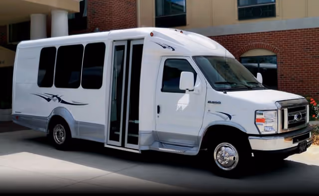 White shuttle bus parked outside a brick senior living facility entrance.