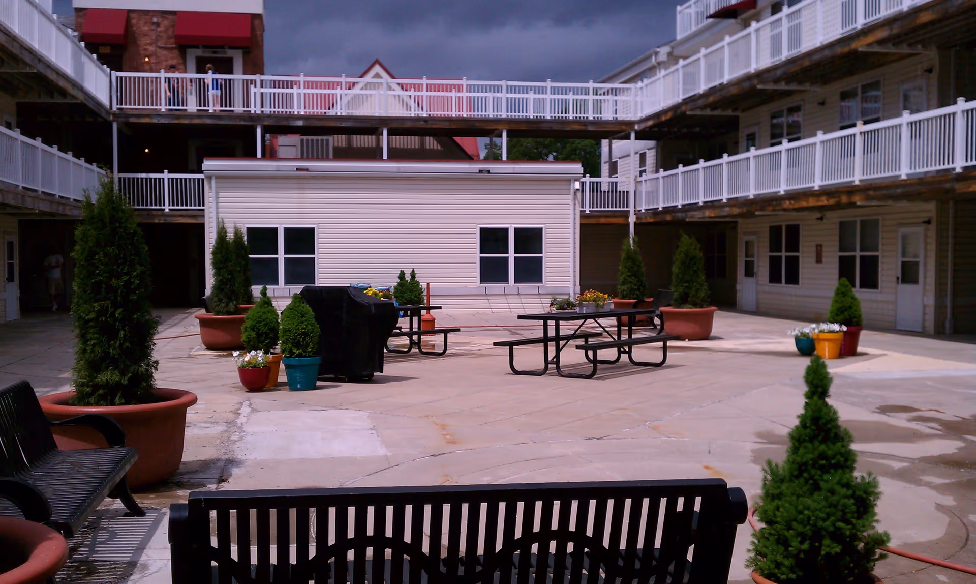 Outdoor courtyard area of Glen Burnie Town Apartments featuring benches, picnic tables, potted plants, and a small building with windows. The courtyard is surrounded by multi-story buildings with white railings and balconies.