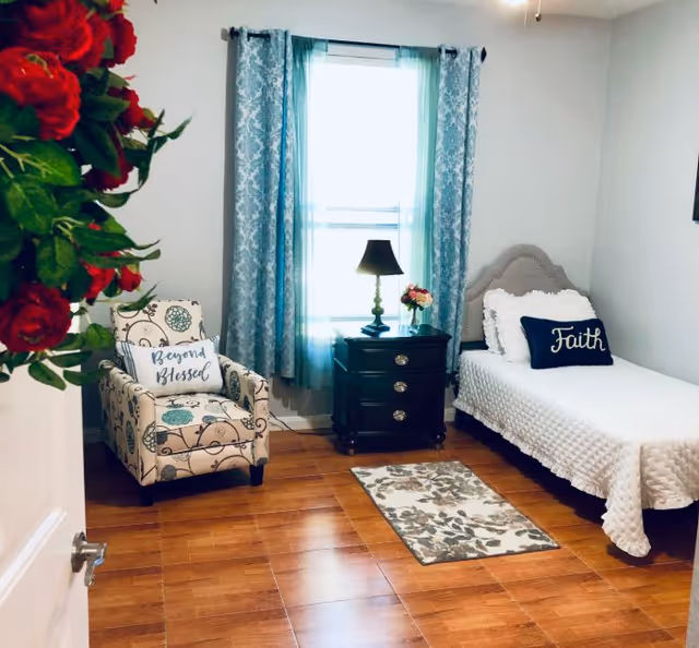 A cozy bedroom at Safe Harbor featuring a single bed, patterned chair, window, and nightstand.