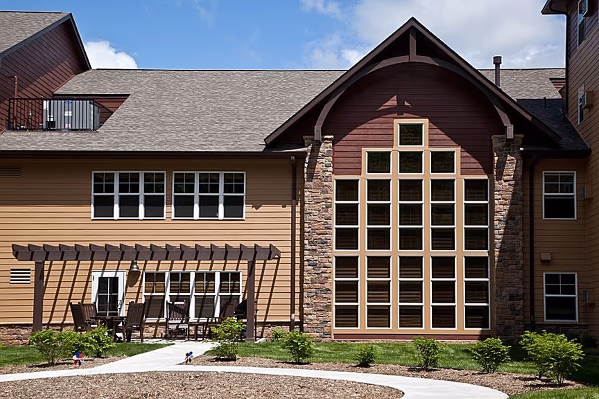 Exterior view of a senior living facility building with beige and brown siding, large multi-pane windows, a stone accent wall, and a small patio area with a pergola and outdoor seating. The sky is blue with some clouds.