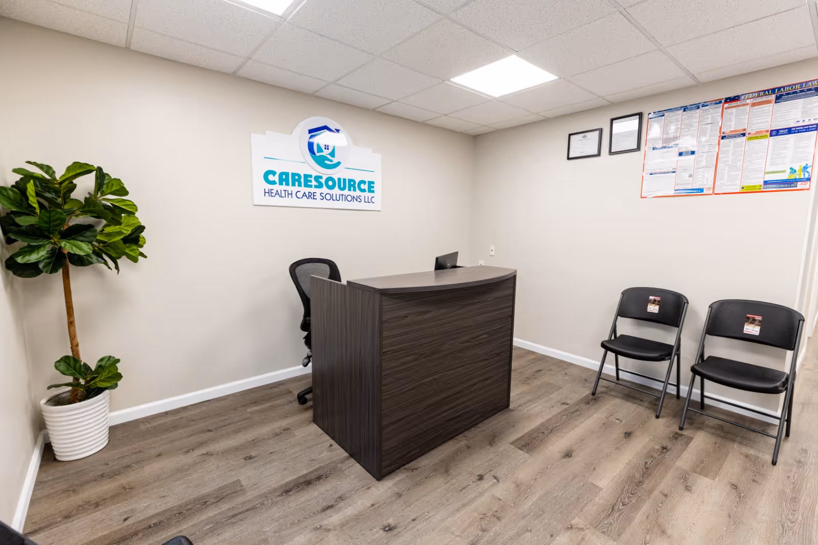 Reception area with a dark wood desk, sign, plant, and two chairs.