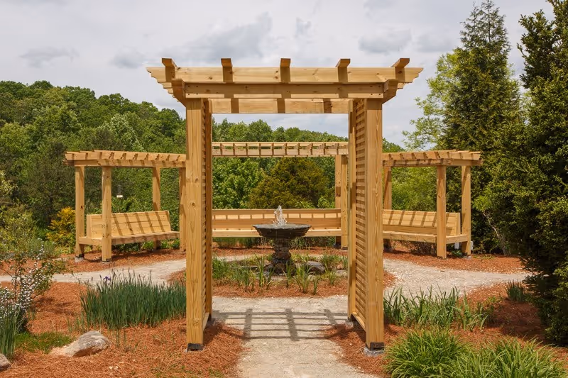Wooden pergolas frame a central fountain with benches in a landscaped outdoor garden.