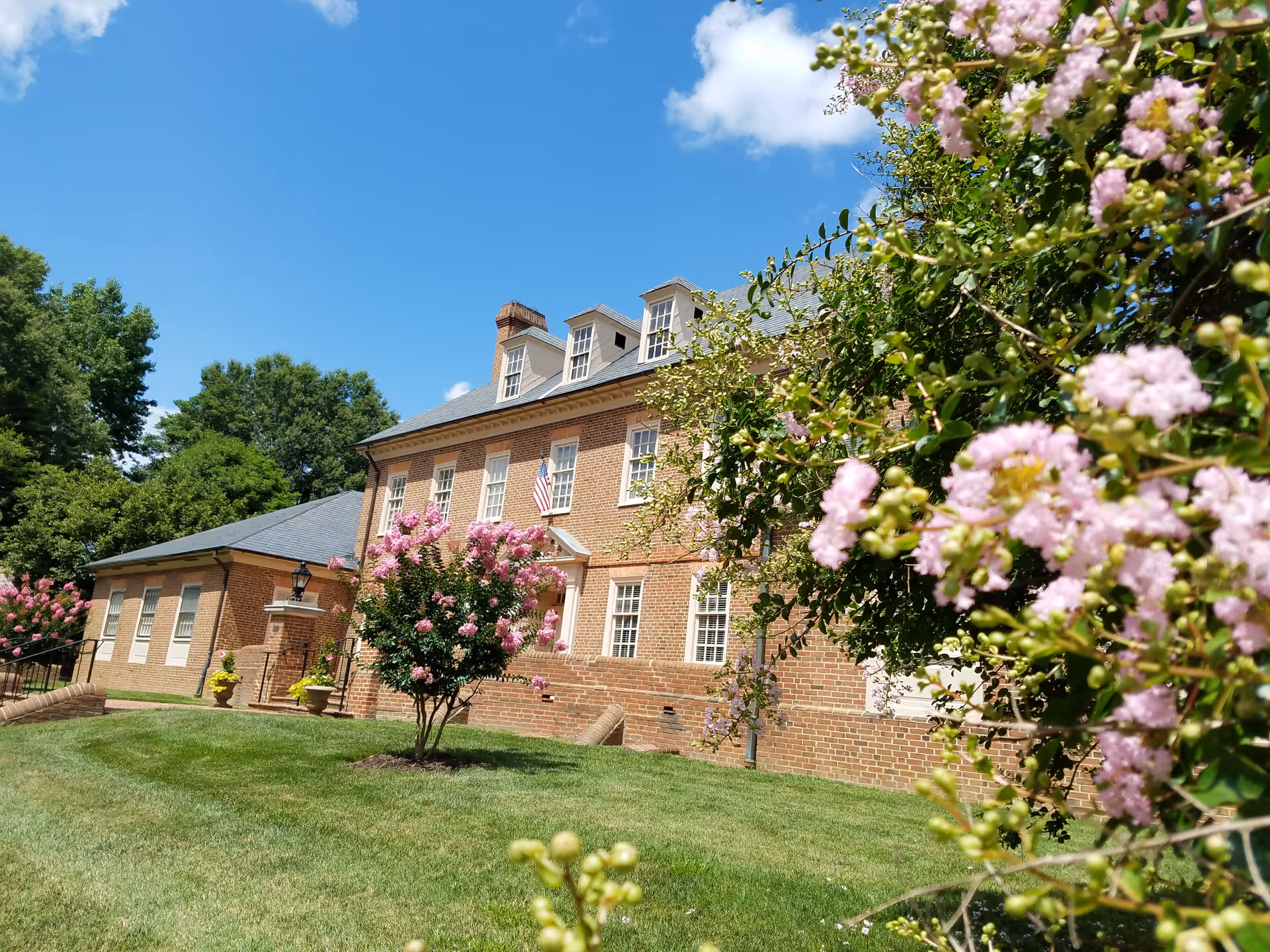 Brick senior living building with green lawn and pink flowering shrubs under blue sky.