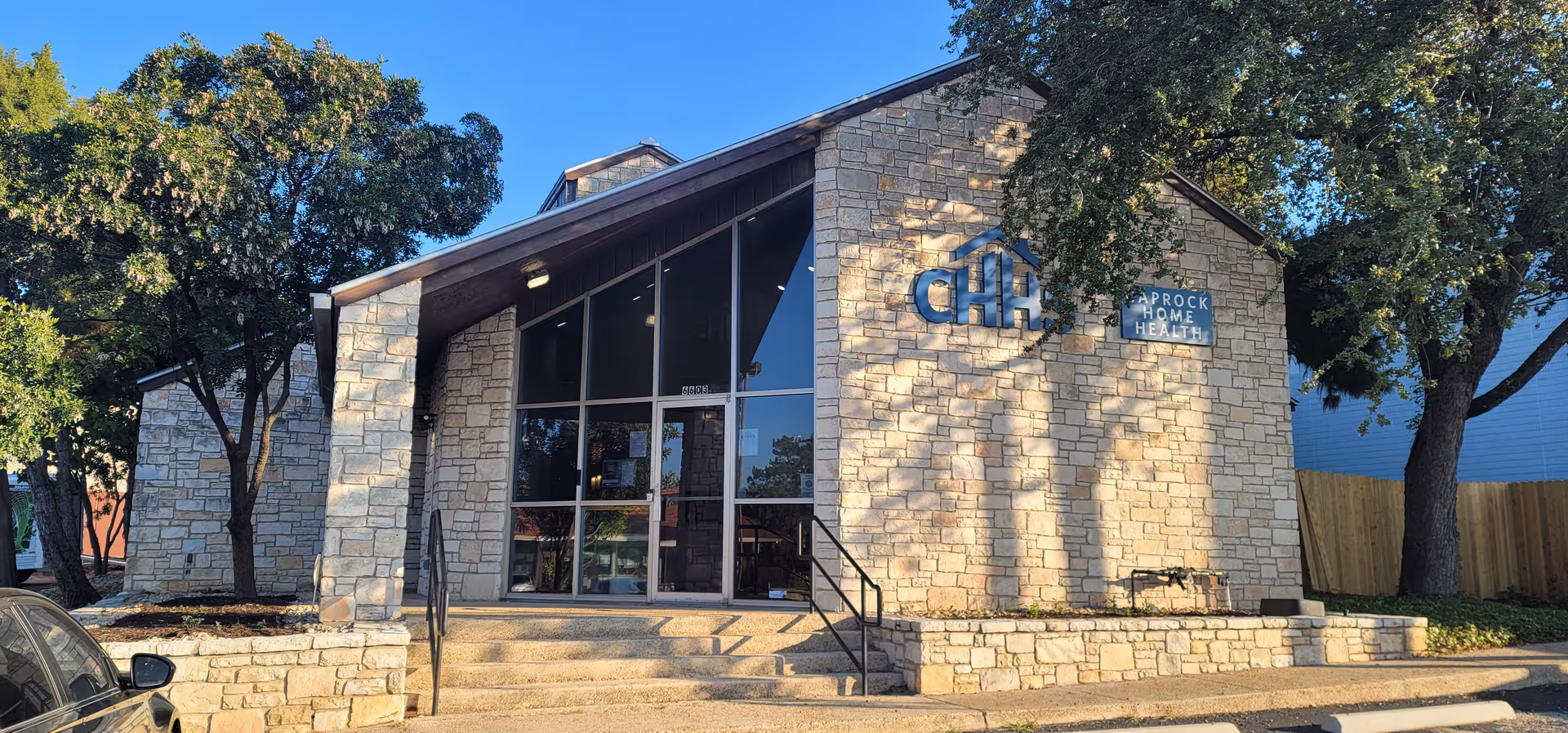 Stone-faced building entrance with large glass doors and Caprock Home Health sign, surrounded by trees.
