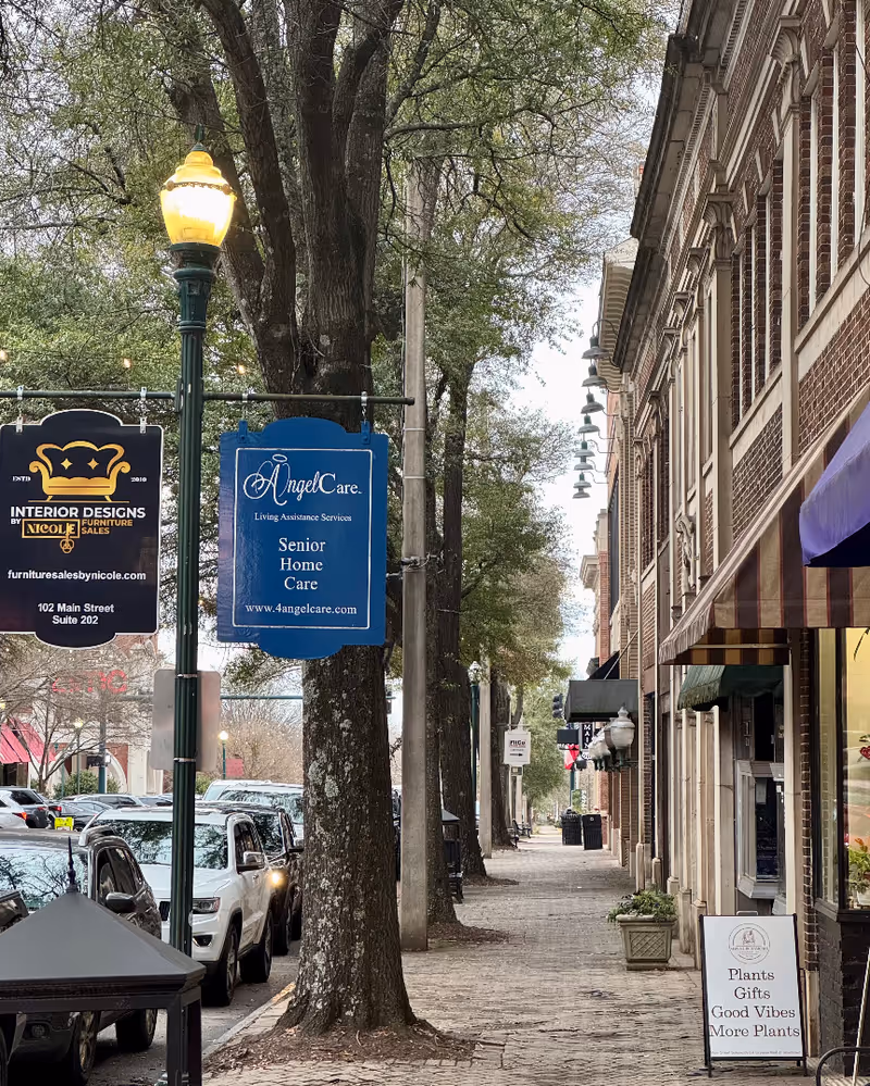 City street view with trees and a blue Angel Care Senior Home Care sign near storefronts