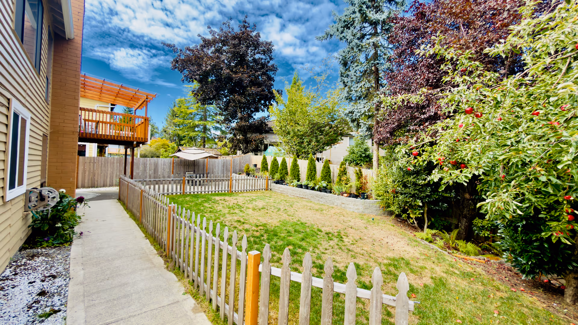 Outdoor fenced backyard with lawn, trees, and seating near a wooden deck.
