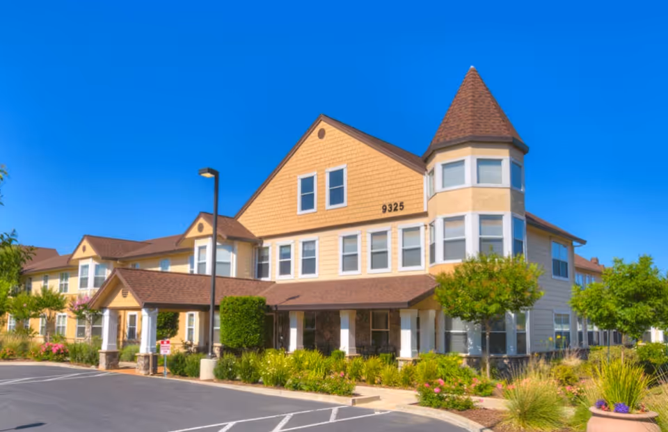 Front view of a two-story yellow senior living building with a turret, landscaped entrance, and parking under a clear blue sky.