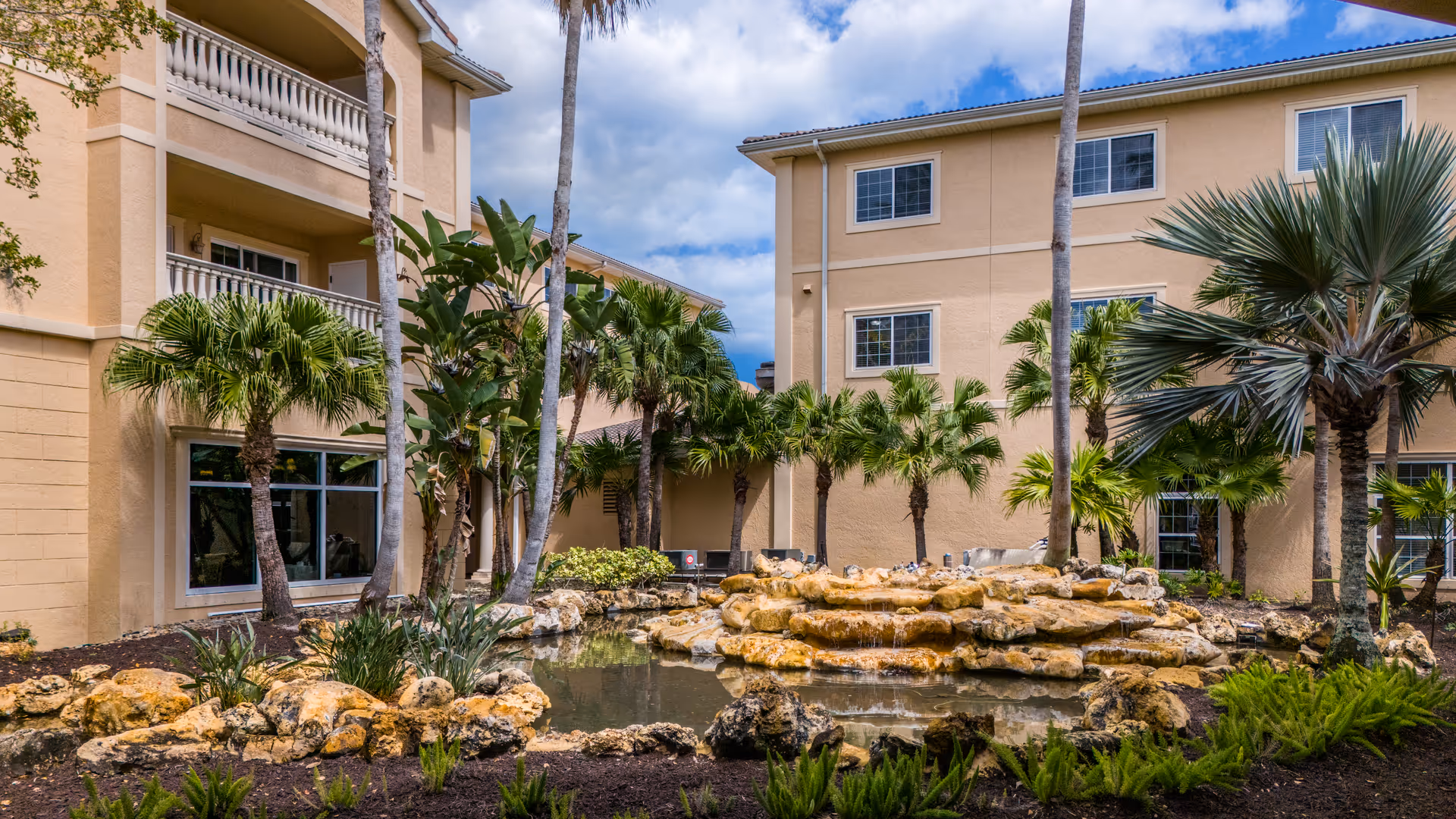 Landscaped courtyard with a pond and rock waterfall in front of a multi-story beige building surrounded by palm trees.