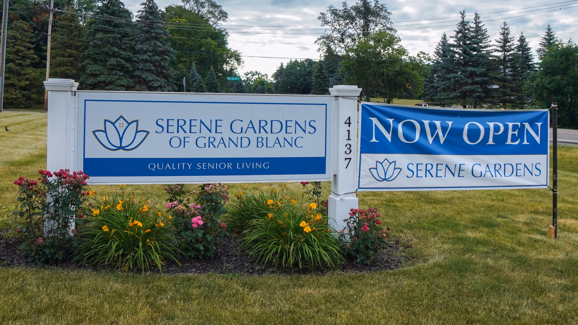 Exterior sign for Serene Gardens of Grand Blanc with a Now Open banner and flowers.