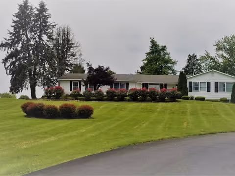 Single-story white building with black shutters surrounded by well-maintained green lawn, bushes, and trees under a cloudy sky.