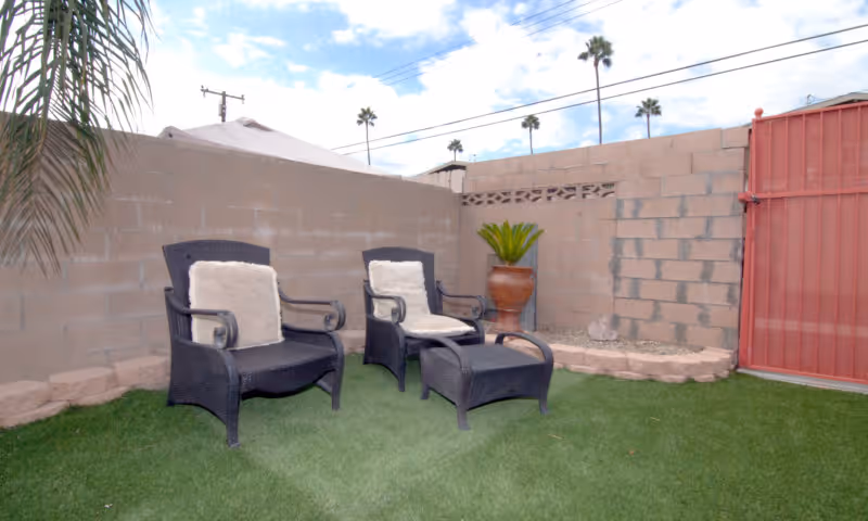 Outdoor patio area with two black wicker chairs with beige cushions and a matching ottoman on green artificial grass, surrounded by a beige brick wall and a large terracotta planter with a green plant. A red metal gate is visible on the right side.