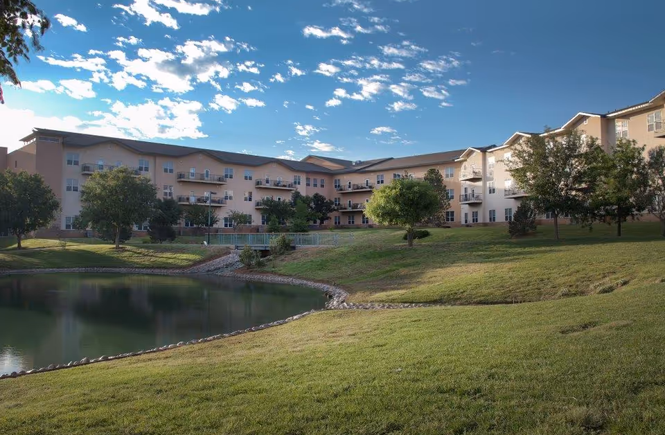 Exterior view of a multi-story senior living building with balconies overlooking a pond and grassy lawn under a partly cloudy sky.