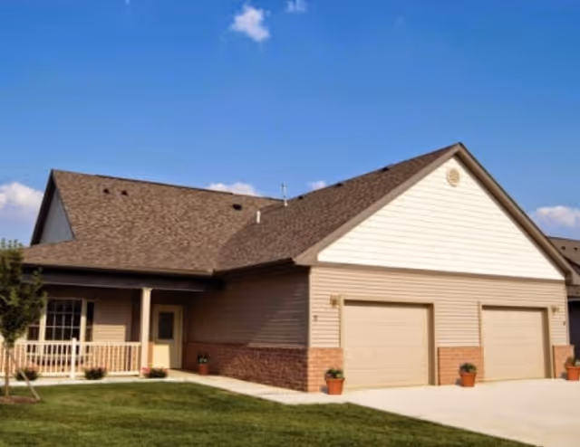 Front exterior of a single-story residential-style building with two garage doors, a small porch, and a manicured lawn under a blue sky.