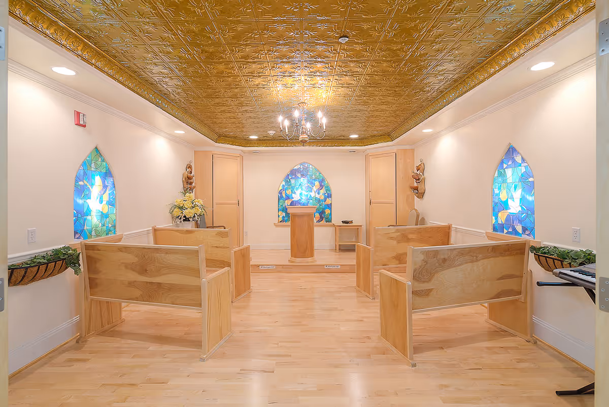 Small chapel-style room with wooden pews, stained glass windows, and a podium beneath an ornate gold ceiling.