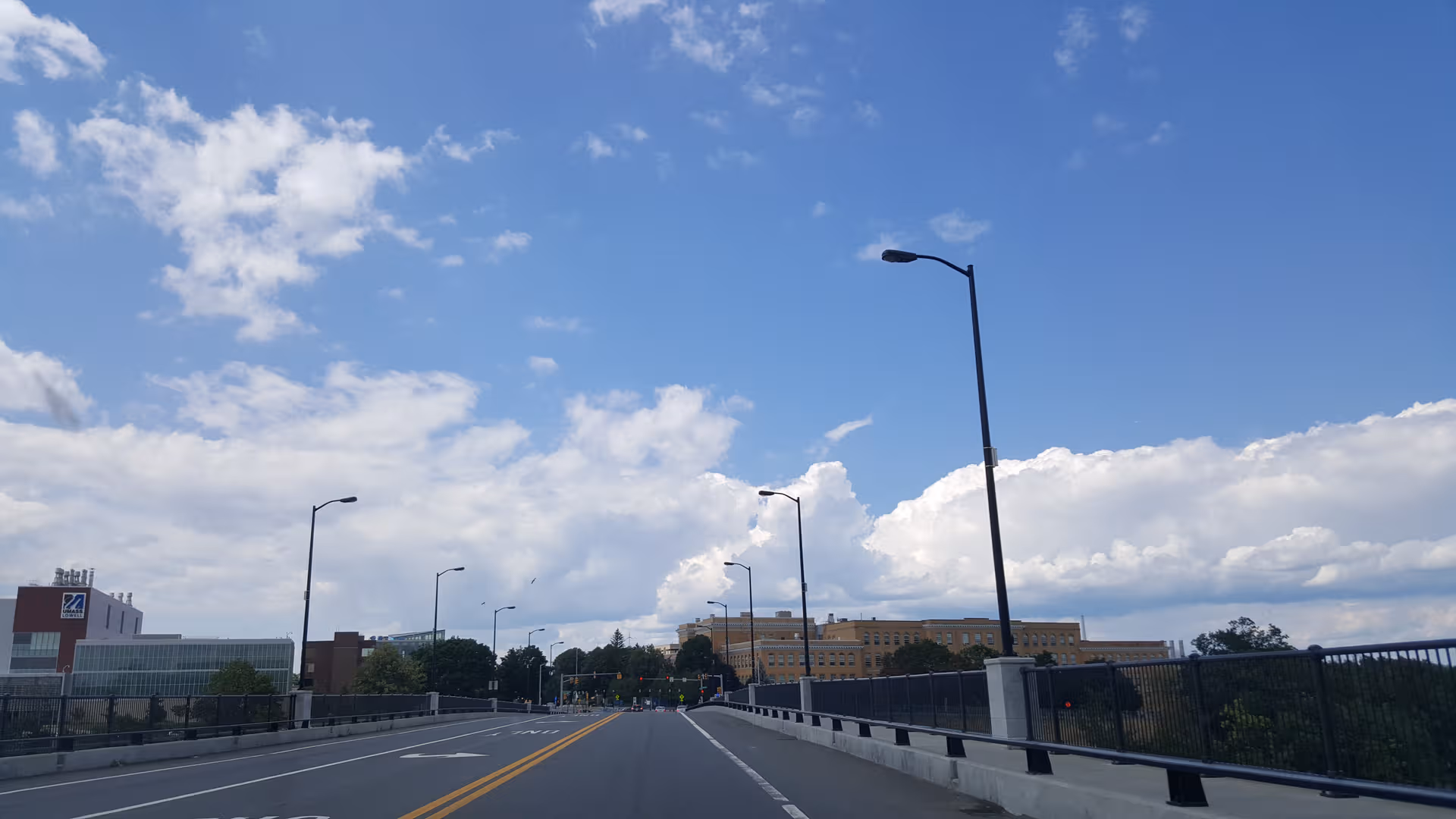 Wide street with streetlights and a railing leads toward a distant large building under blue sky.