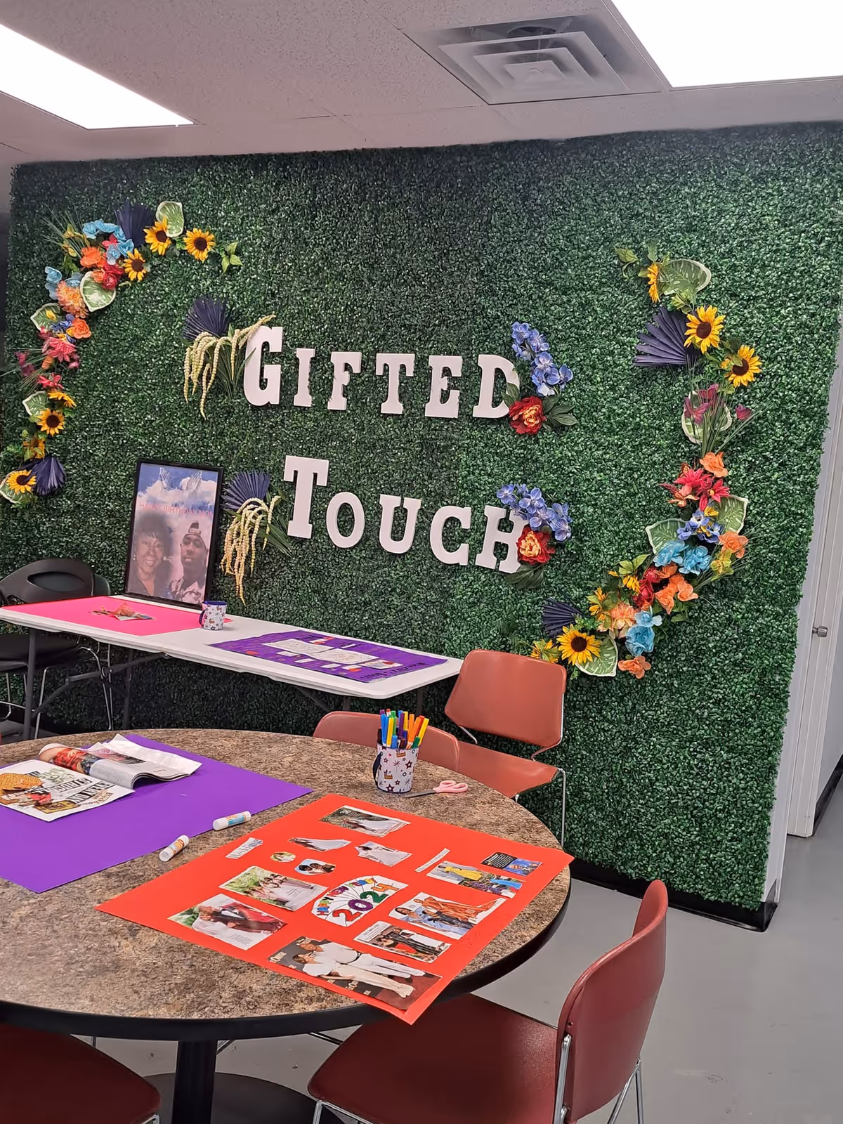 Green hedge wall with Gifted Touch sign and colorful craft tables.