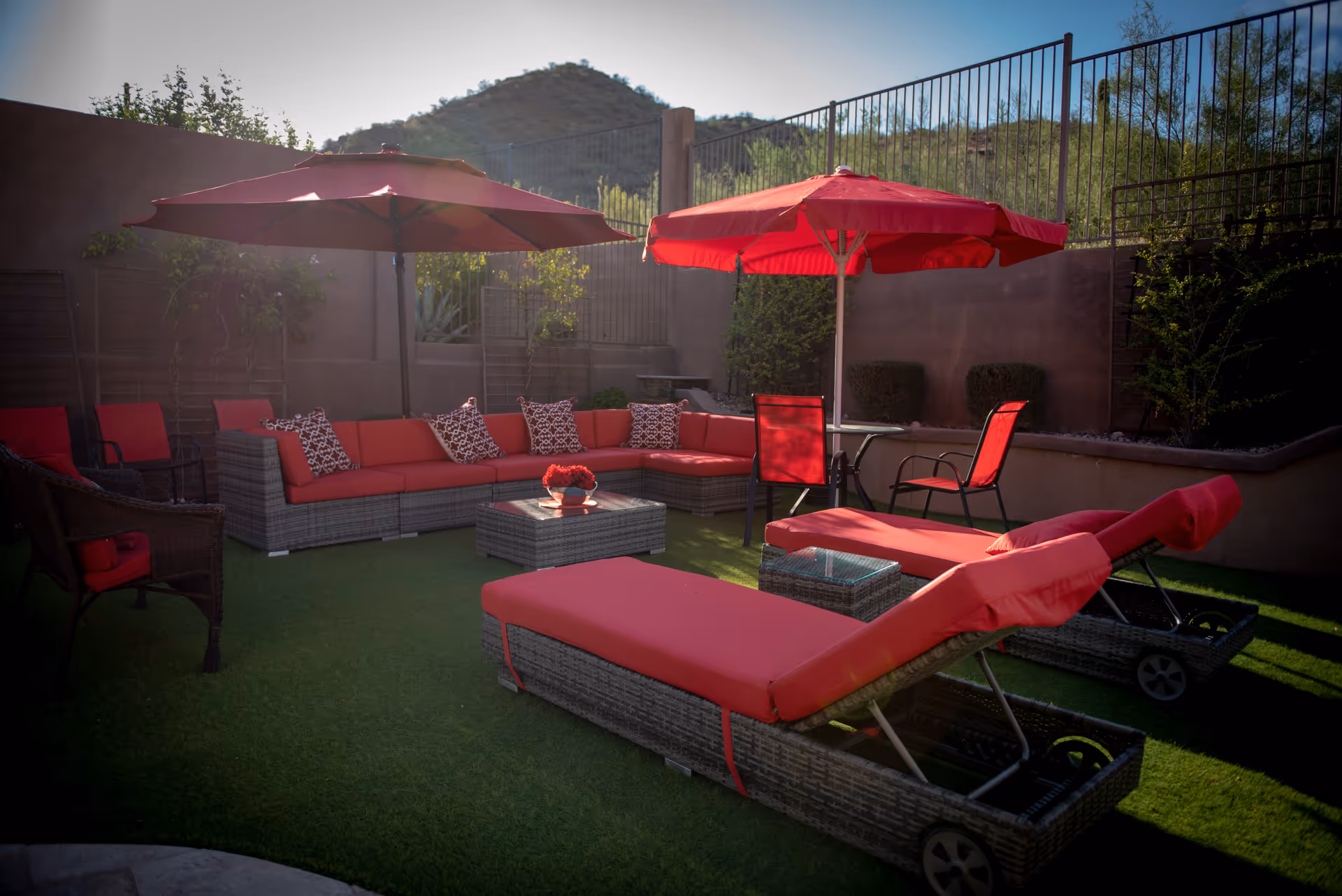 Outdoor patio area with red-cushioned lounge chairs, a sectional sofa, umbrellas, and a coffee table on artificial grass by a fenced wall.