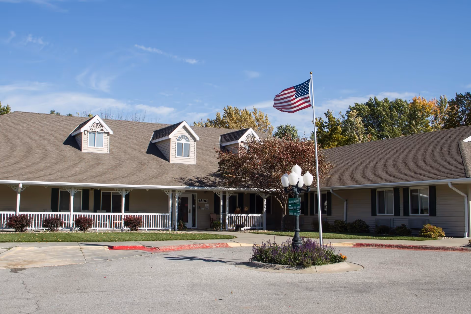 Front exterior of a single-story senior living facility with a covered porch, American flag on a pole, and circular driveway.