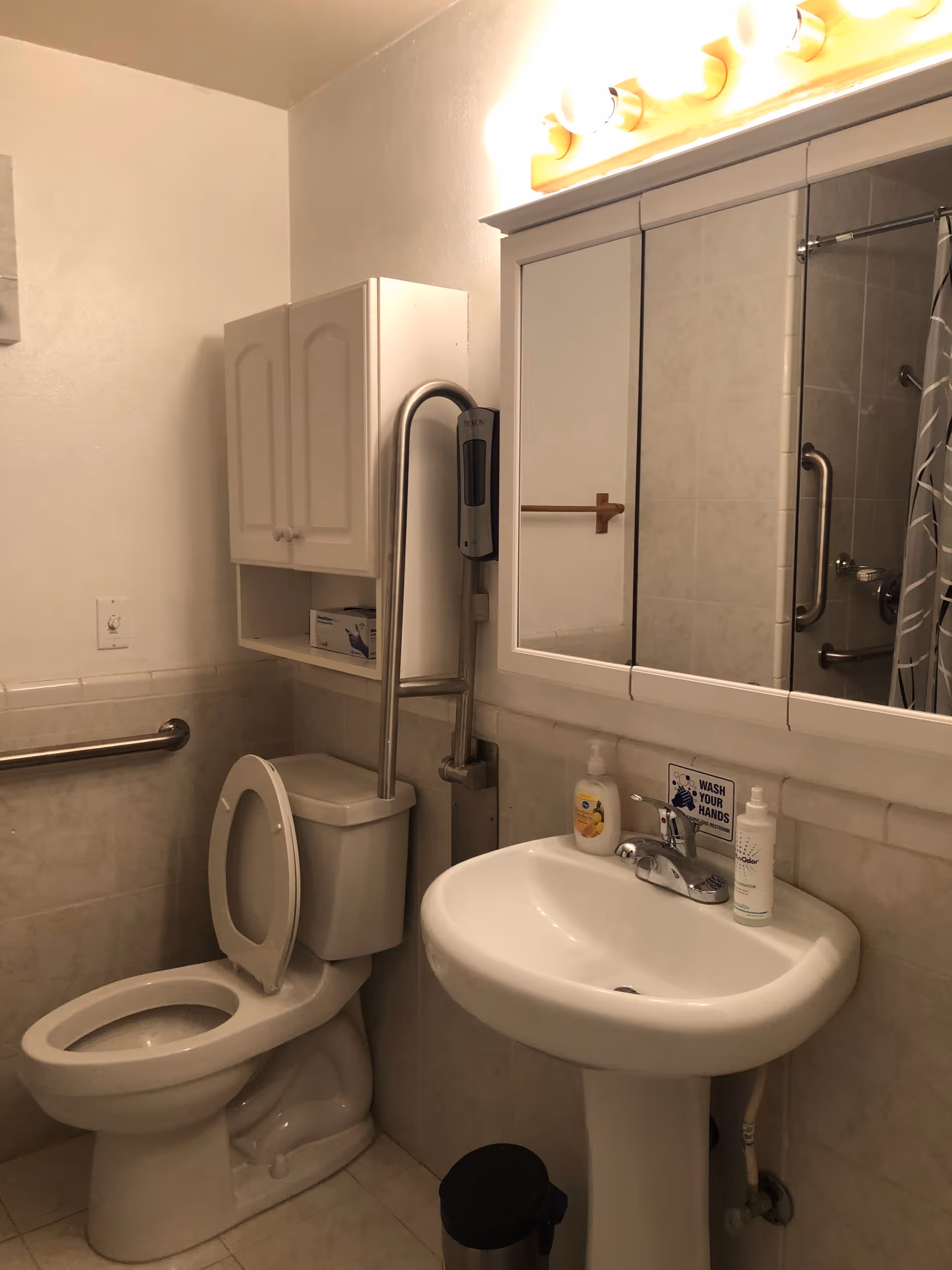 Accessible bathroom in a senior living facility with toilet, sink, mirror cabinet, grab bars, and shower.