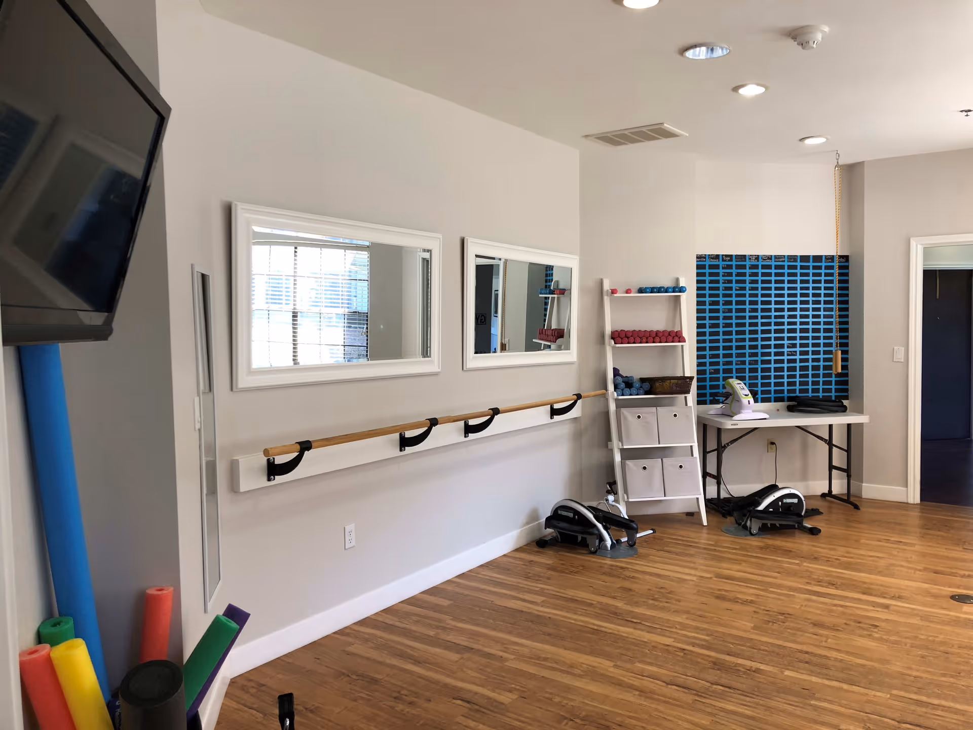 A fitness or therapy room with wooden floors, two wall-mounted mirrors, a wooden handrail, a white shelving unit holding small dumbbells and exercise equipment, and a table with additional exercise devices. Colorful foam rollers are visible in the corner, and a large TV is mounted on the wall.
