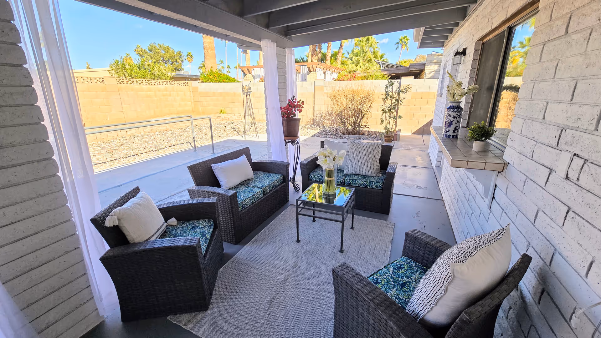 Covered outdoor patio with wicker seating, a glass table, and potted plants along brick walls.