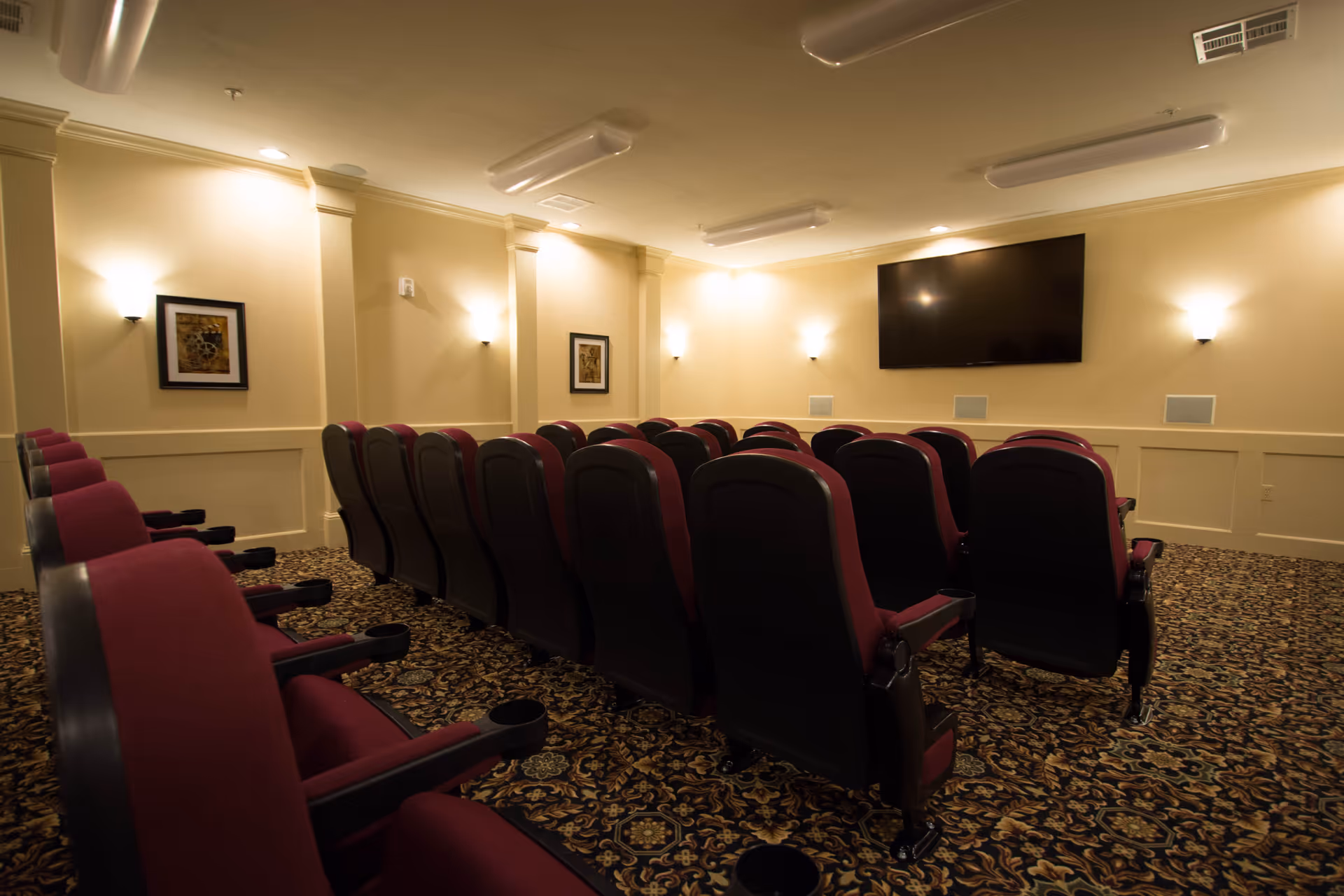 Small interior theater room with rows of red upholstered seats facing a wall-mounted television.