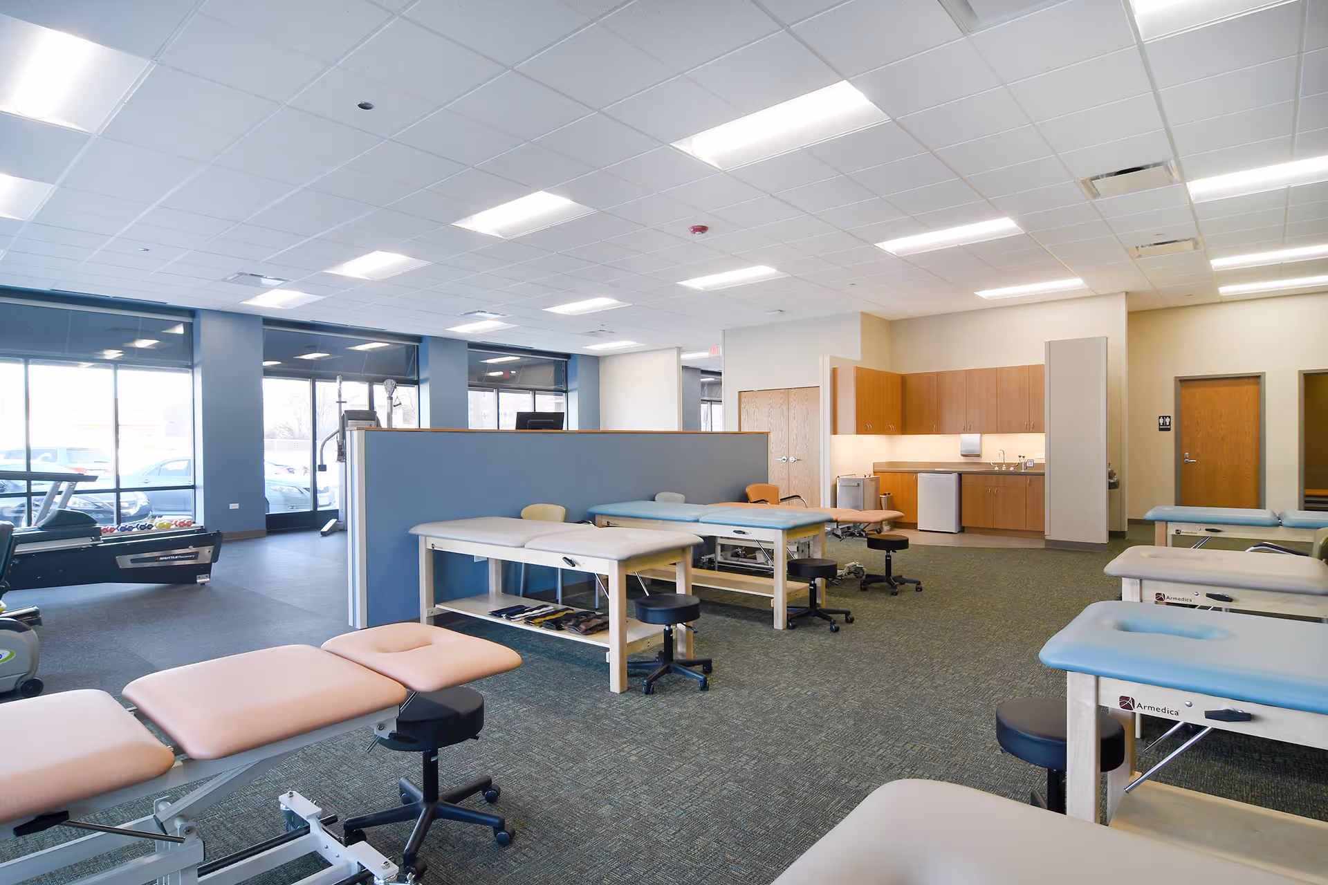 A bright, open physical therapy room with treatment tables and a kitchenette.