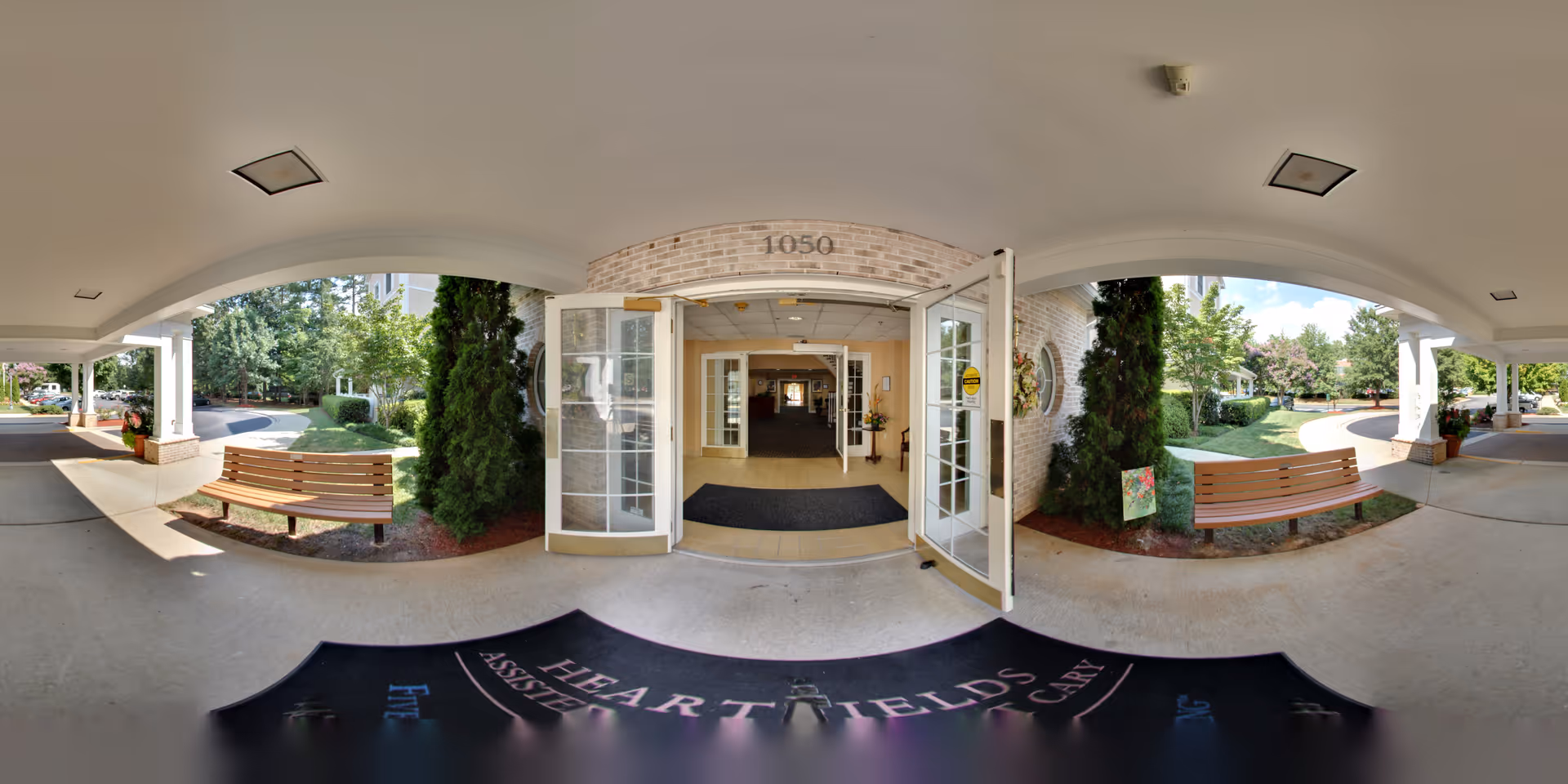 Entrance area of HeartFields at Cary facility showing double glass doors open to an interior hallway. Outside the entrance, there are two wooden benches on either side with greenery and trees around. The building number 1050 is displayed above the door. The area is covered by a white ceiling with recessed lighting.