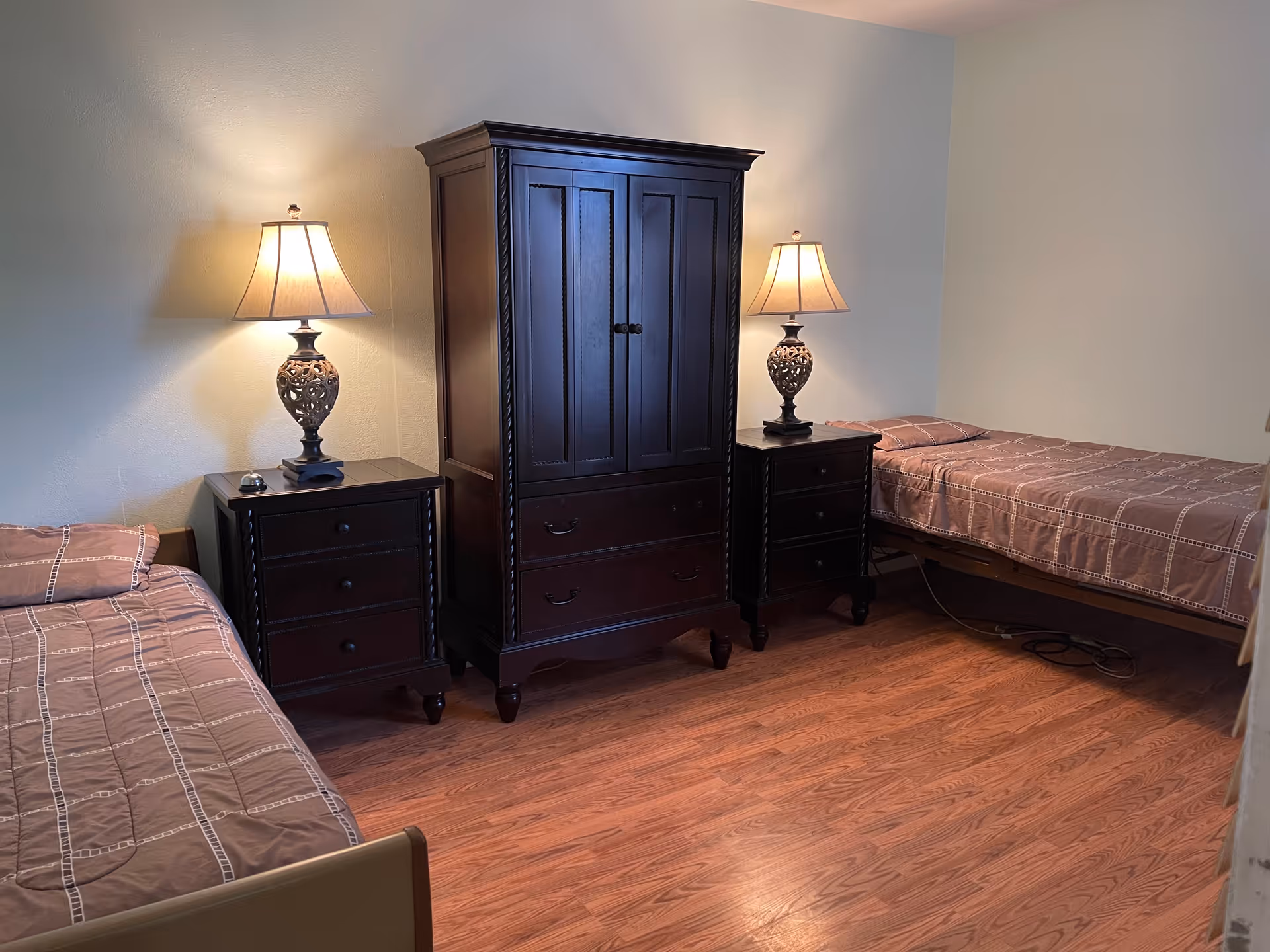 A bedroom with two single beds on opposite sides of the room, each with a brown patterned bedspread. Between the beds is a dark wooden armoire flanked by two matching nightstands, each topped with a decorative lamp emitting warm light. The floor is wooden, and the walls are painted a light color.
