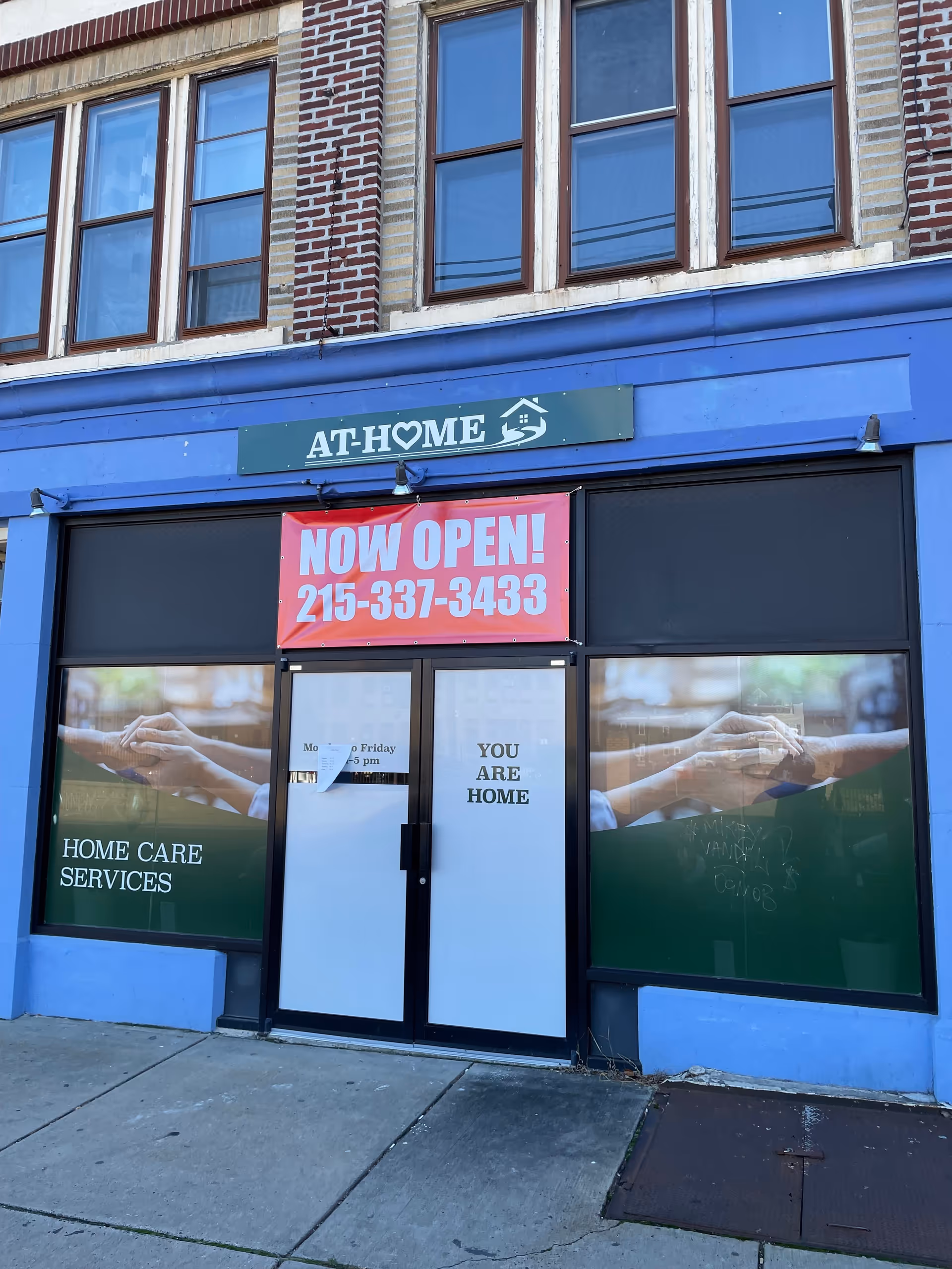 Blue storefront with AT-HOME sign and NOW OPEN banner for a home care services facility.