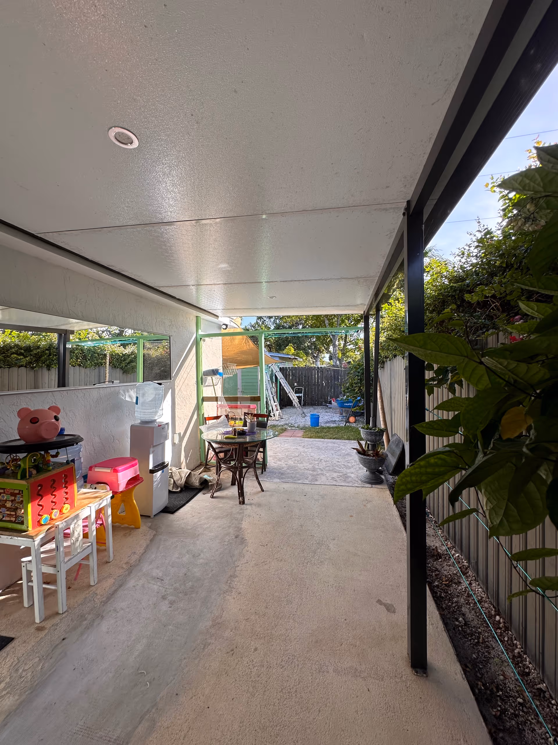 Covered outdoor patio area with a concrete floor, a small table with chairs, a water dispenser, children's toys including a pink pig toy and a colorful activity cube, and plants along the right side near a metal fence. In the background, there is a yard with grass, a wooden fence, and some outdoor items like a ladder and a blue bucket.