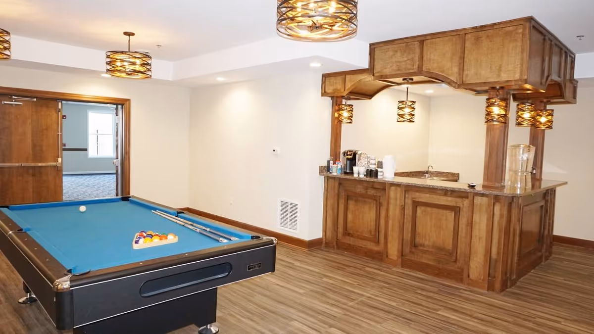 A recreational room featuring a blue felt pool table with pool cues and balls arranged for a game. To the right, there is a wooden bar area with hanging pendant lights, a coffee maker, cups, and a water dispenser. The room has wood flooring and white walls with recessed lighting.