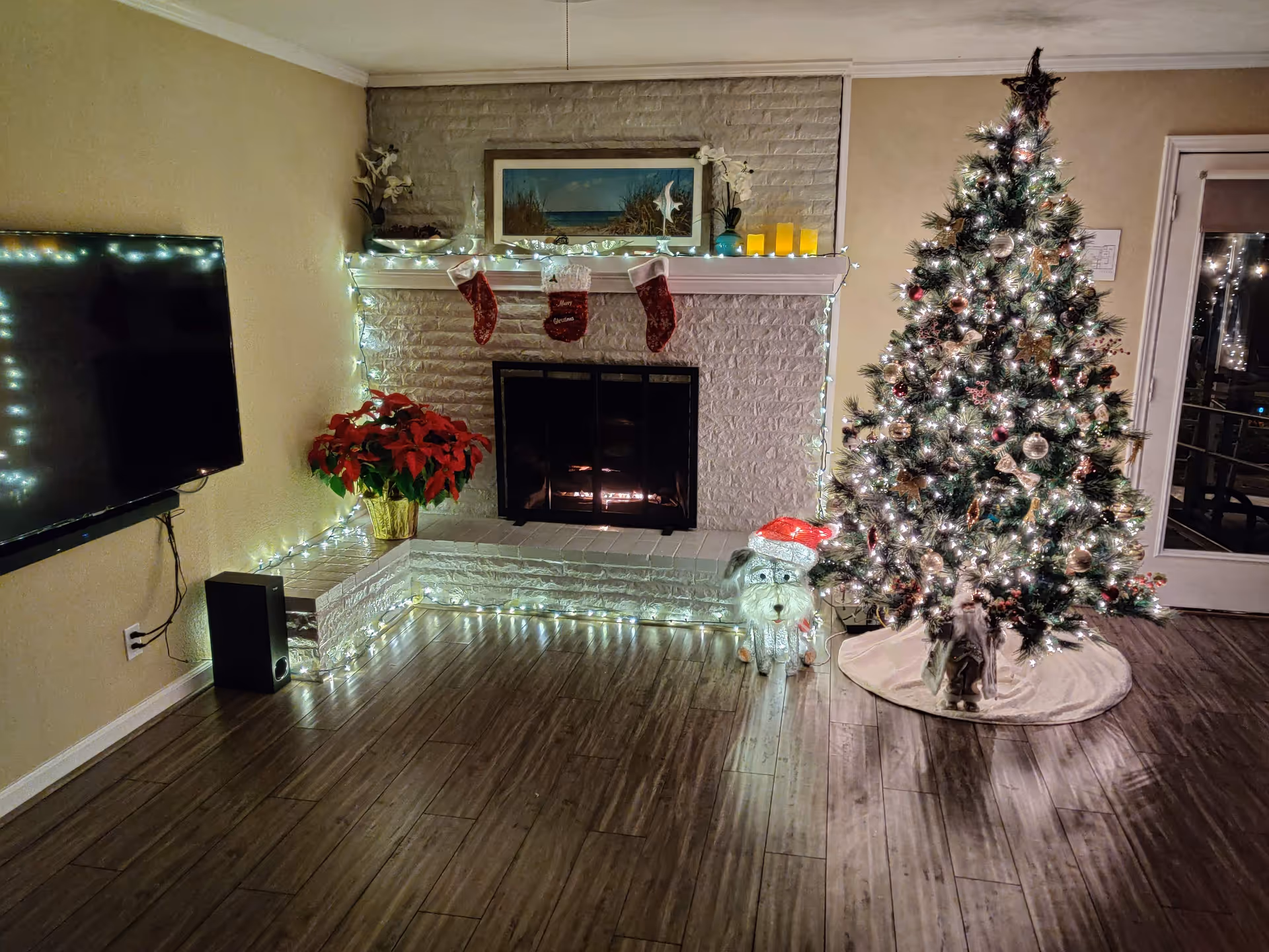 Cozy living room with lit fireplace, Christmas tree, stockings, twinkling lights, poinsettia, and TV.