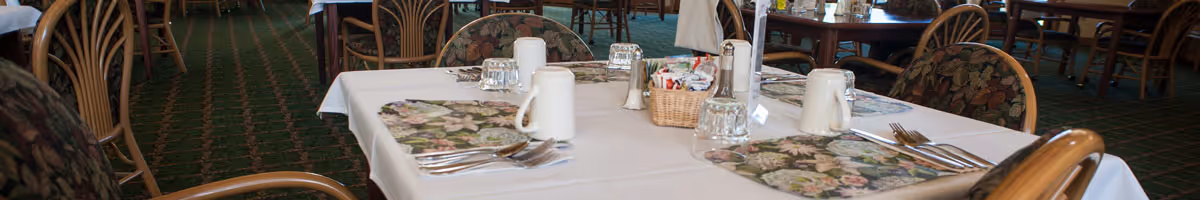 A communal dining room table set with floral placemats, silverware, pitchers, glasses, and cushioned chairs.