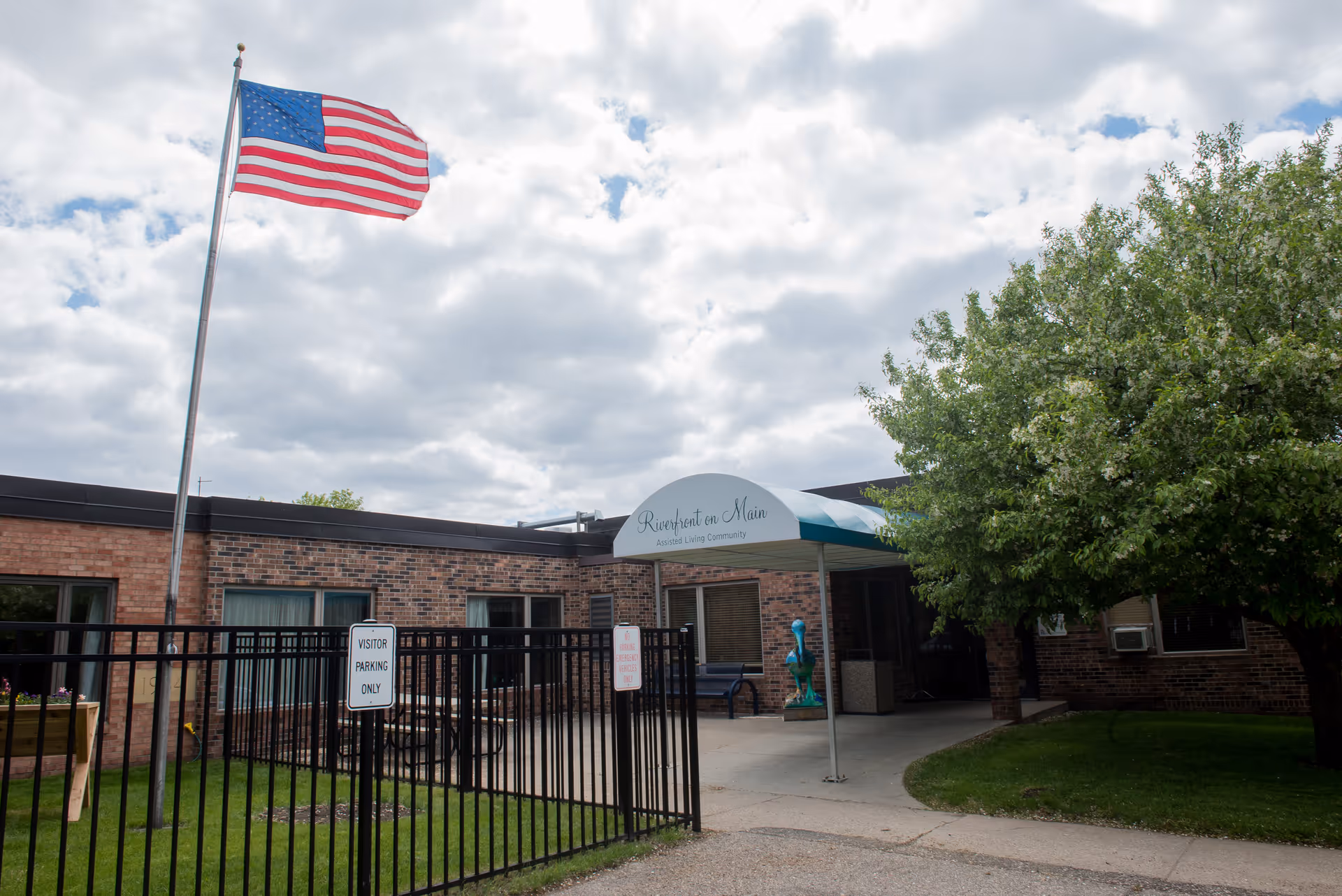 Flag waves beside a brick senior living center entrance with a curved canopy.