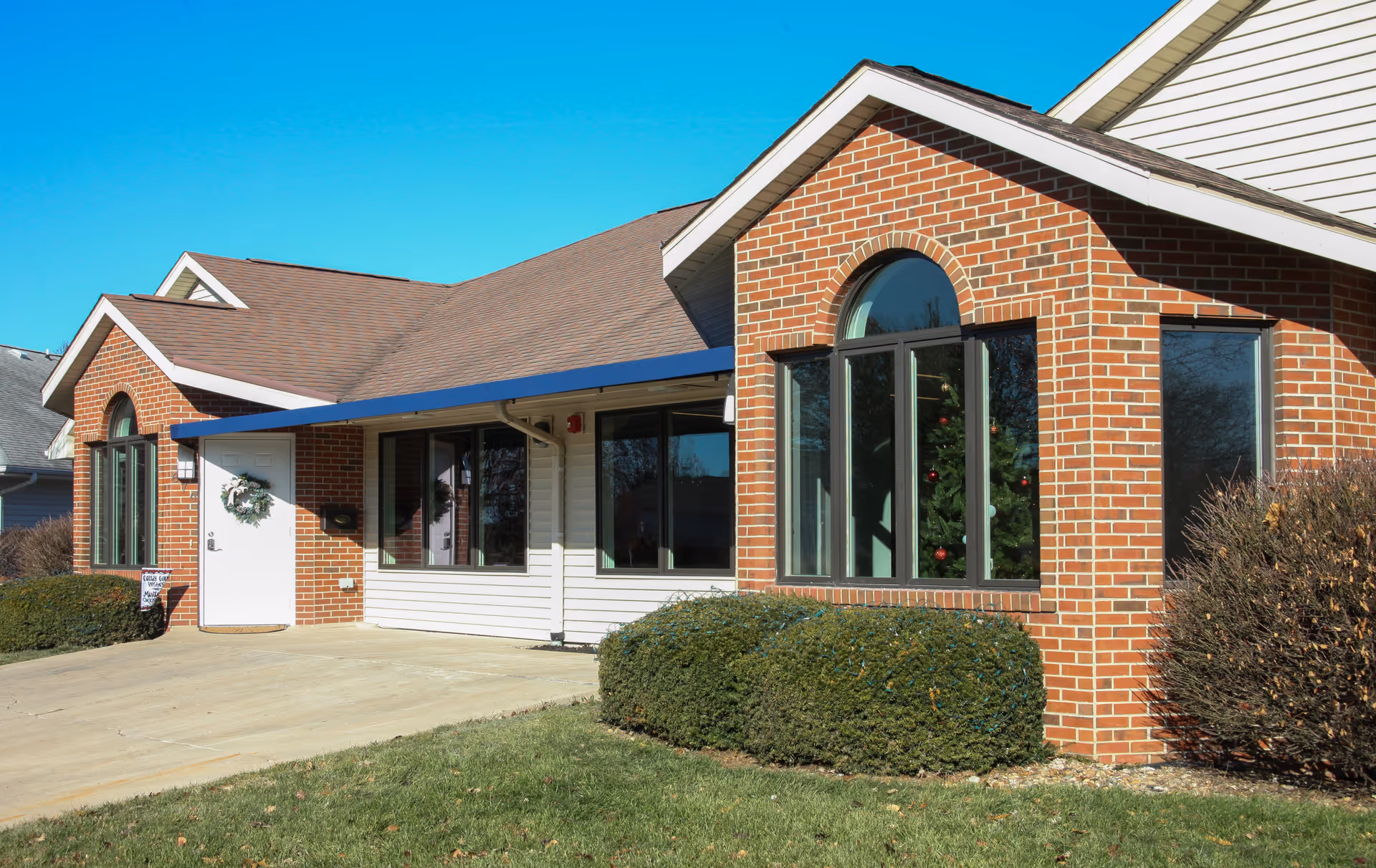 Front exterior of a single-story brick and siding building with arched windows, a white door with a wreath, and landscaped shrubs.
