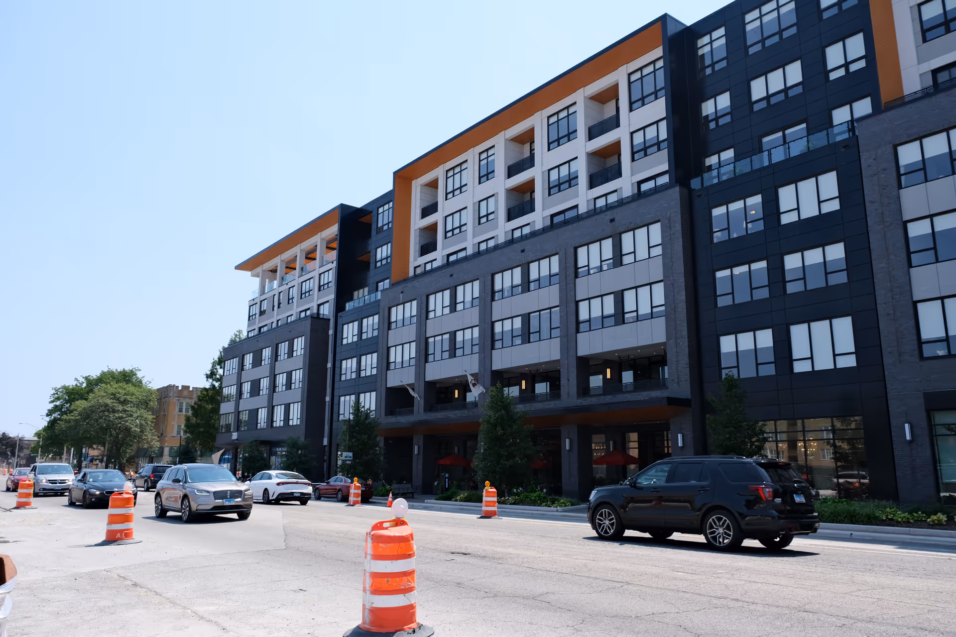 Street view of a modern multi-story residential building with cars and orange construction cones in front.