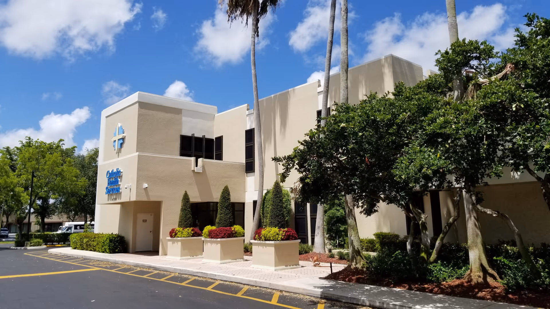 Exterior view of a beige Catholic Health Services building with landscaping and palm trees.