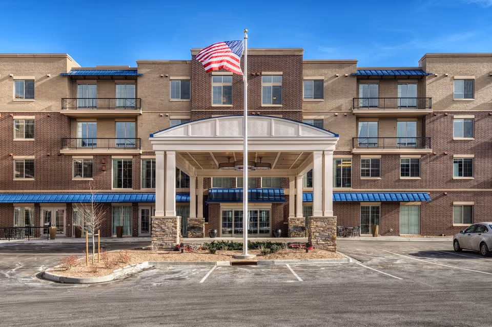 Three-story brick senior living facility with a covered entrance, flagpole, and parking lot.